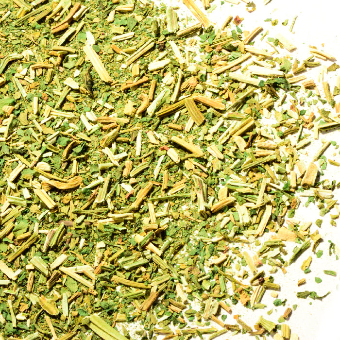 A close-up of Whole Spice Inc. Epazote Leaves Crushed scattered on a white surface, showing green leaves and tiny yellowish stems—ideal for adding to herbal tea infusions.