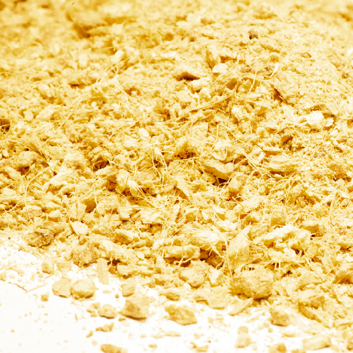 A close-up of a scattered pile of Whole Spice Inc. Ginger Crushed, showing its vibrant yellow flakes and powder, resembling a spice blend or curry powder, on a white surface.