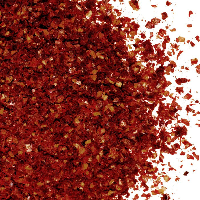 A close-up of Harissa Spice Coarse by Whole Spice Inc. shows small, irregular red and yellowish flakes of this spicy seasoning scattered on a white background.
