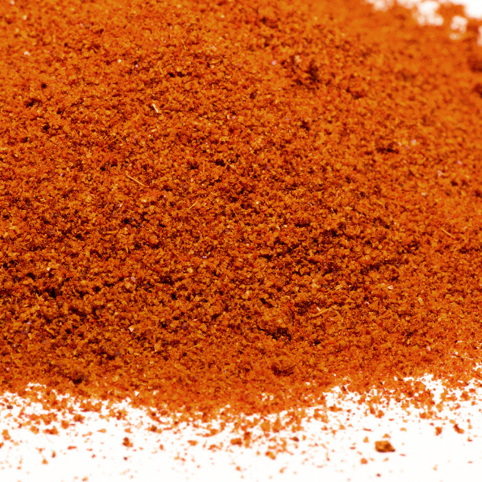 A close-up of Harissa Spice by Whole Spice Inc., showing a vibrant reddish-orange, finely ground blend on a white surface, highlighting its bold color and texture.