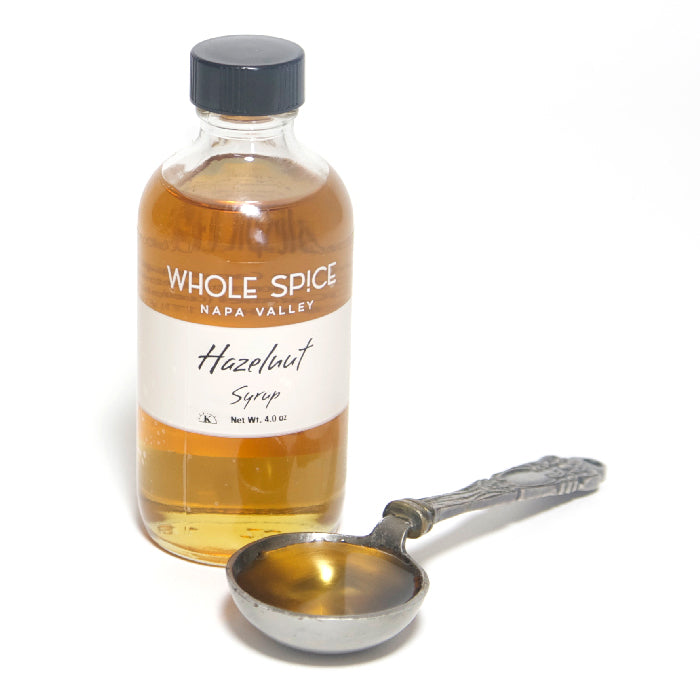 A glass bottle labeled Whole Spice Inc. Hazelnut Syrup stands next to an ornate metal spoon filled with amber syrup, ideal for adding rich hazelnut flavor to your favorite coffee drinks.