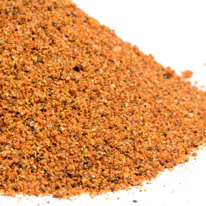 A close-up of Whole Spice Inc. Honey BBQ Rub shows a mound of reddish-brown, finely ground seasoning with flecks of black, orange, and yellow spices on a white background.