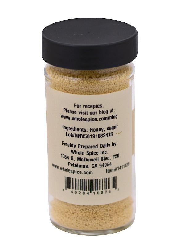A clear glass jar from Whole Spice Inc., labeled "Honey Granulated," has a black lid and contains light brown honey granules. The label lists honey and sugar as ingredients, manufacturer info, website, and a visible barcode.