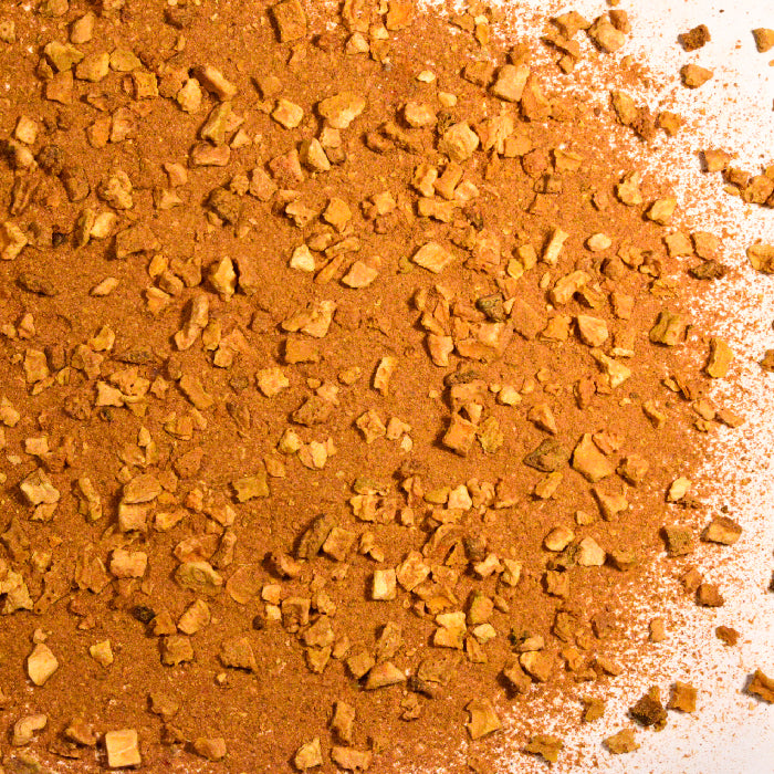 A close-up of Whole Spice Inc. Hot Orange Tea Caffeine Free shows a reddish-brown blend with fine powder and chunky orange peel pieces, spread on a white surface.