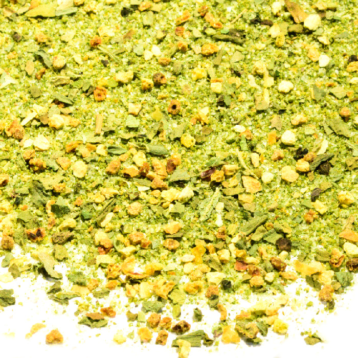 Close-up of Whole Spice Inc. Key West Style Seasoning—a blend of dried green and yellow herbs and spices, ideal for chicken or fish—scattered in small, textured pieces on a white background.