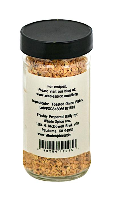 A clear glass jar with a black lid contains Whole Spice Inc. Onion Toasted Flakes, ideal for soup bases. The label lists ingredients, preparation details, dehydrated onion content, a website, and a barcode on the back.