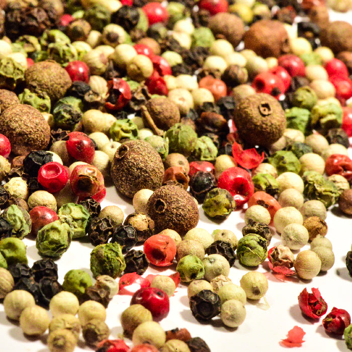 A close-up of Whole Spice Inc. Peppercorns Five Mix shows colorful black, white, green, and red whole peppercorns scattered on a white surface.