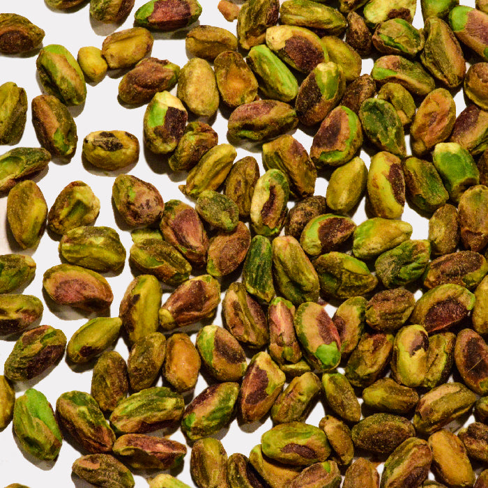 A close-up of Whole Spice Inc. Roasted Pistachios Unsalted scattered on a white background highlights the green, purple, and brown kernels—delicious and healthy for snacking.