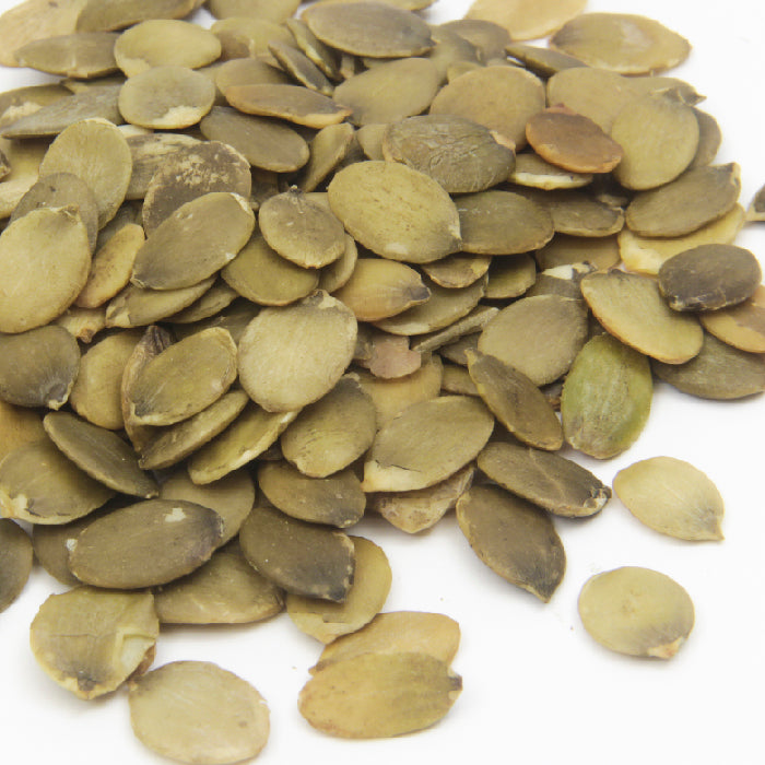 Whole Spice Inc. Pumpkin Seeds Roasted are oval, flat seeds with a greenish-brown color, scattered on a white surface and lightly dusted with sea salt for enhanced flavor.