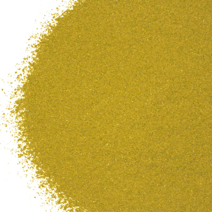 A close-up of finely ground, yellowish-green Sassafras Powder by Whole Spice Inc., spread on a white background. The powder forms a dense, textured area on the right with scattered grains along the left edge.