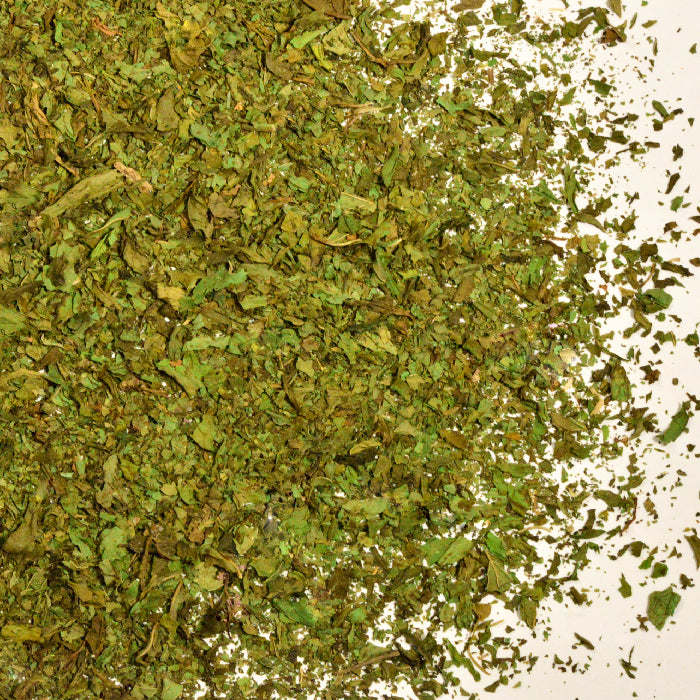 A close-up of Whole Spice Inc. Spearmint Leaves, dried and crushed, scattered on a white surface.