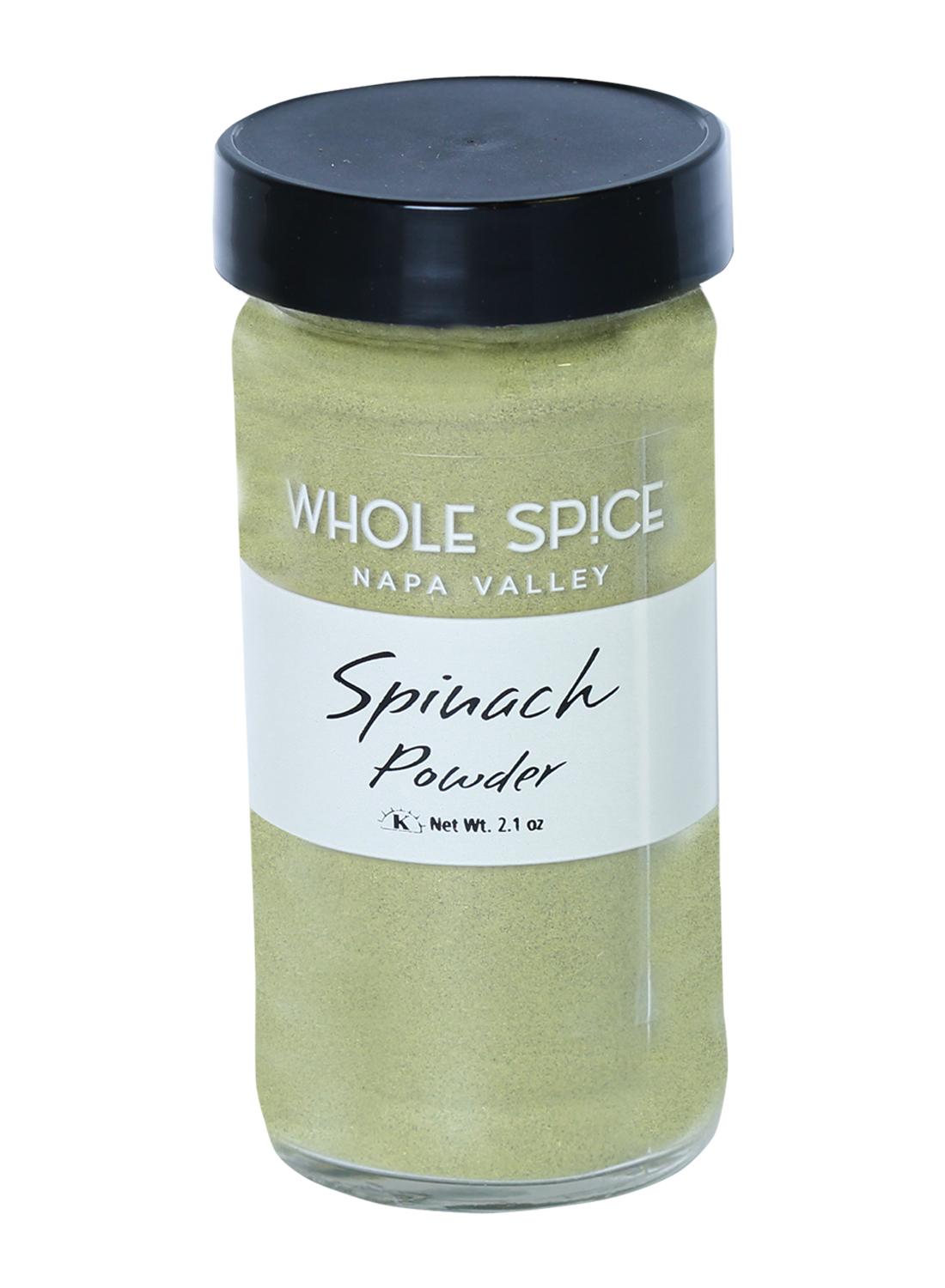 A clear glass jar with a black lid holds green spinach powder. The white label shows Whole Spice Inc. Spinach Powder and lists a net weight of 2.1 oz.