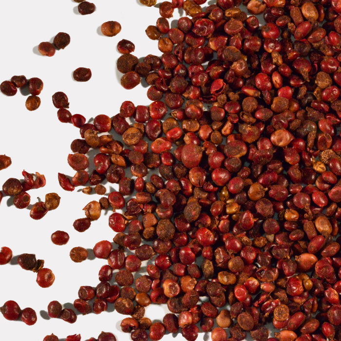 Sumac Whole berries by Whole Spice Inc. and reddish-brown Sichuan peppercorns are scattered on a white surface, with some loose seeds spread out from the main cluster.