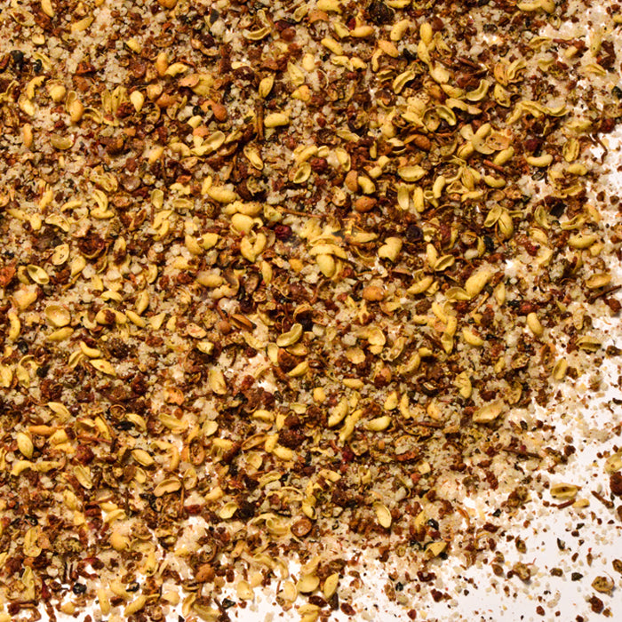 A close-up of Whole Spice Inc. Szechuan Salt, highlighting its coarse brown, yellow, and tan granules and seeds, spread across a white surface.